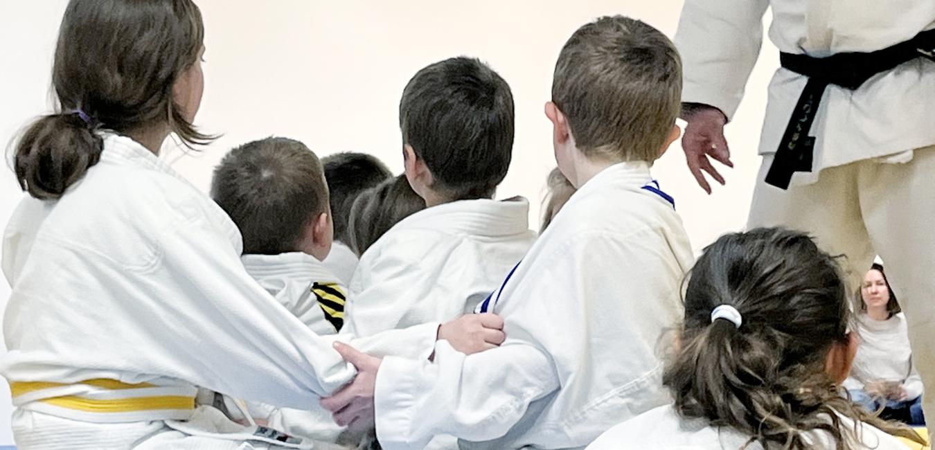 Kinder sitzen in Judoanzügen auf dem Boden und schauen zu ihrem Trainer