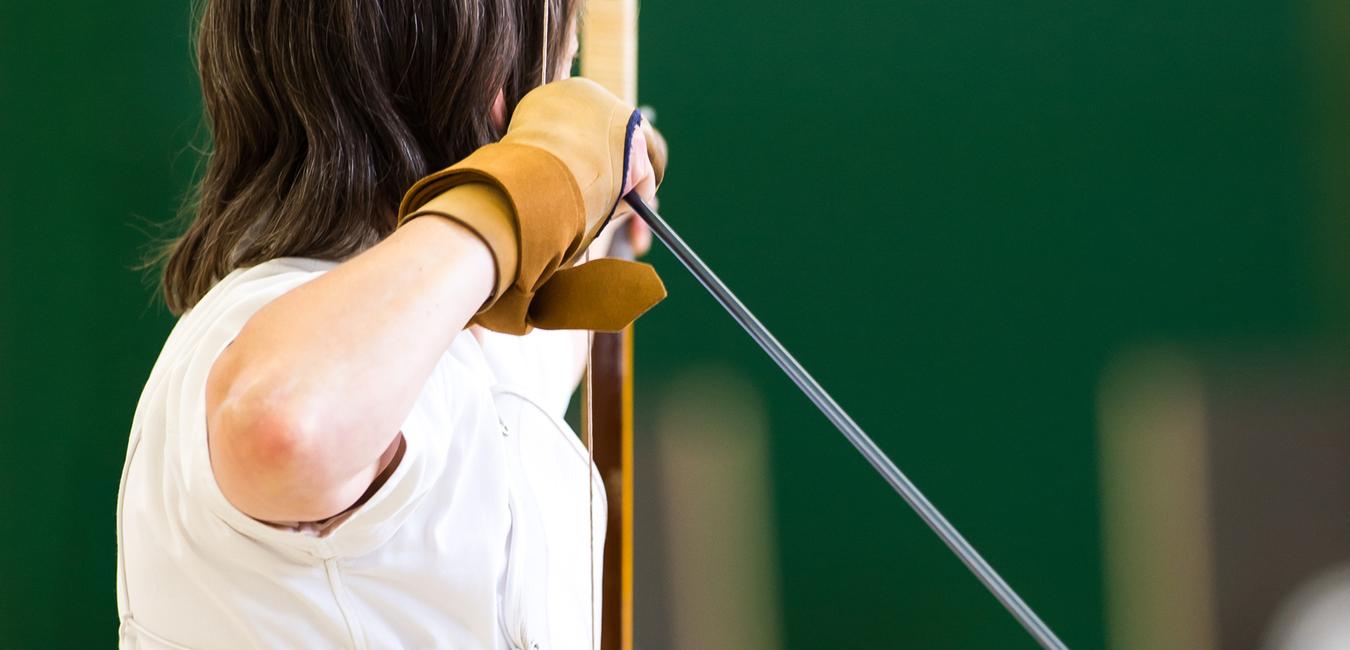 Eine Person in weißem Oberteil und schwarzem Rock zielt mit einem Bogen auf eine Zielscheibe in einer Sporthalle.