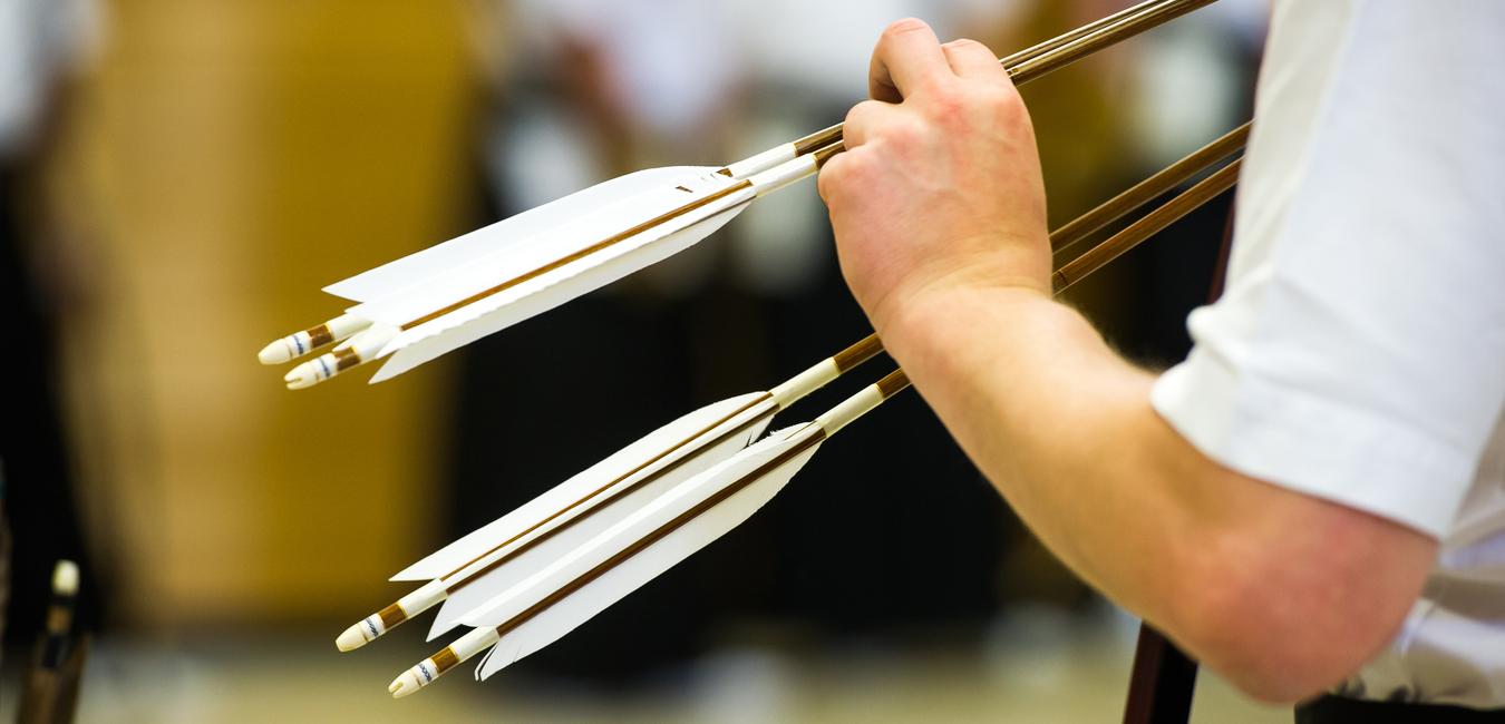 Nahaufnahme einer Hand, die mehrere Pfeile mit weißen Federn in einer Sportumgebung hält.
