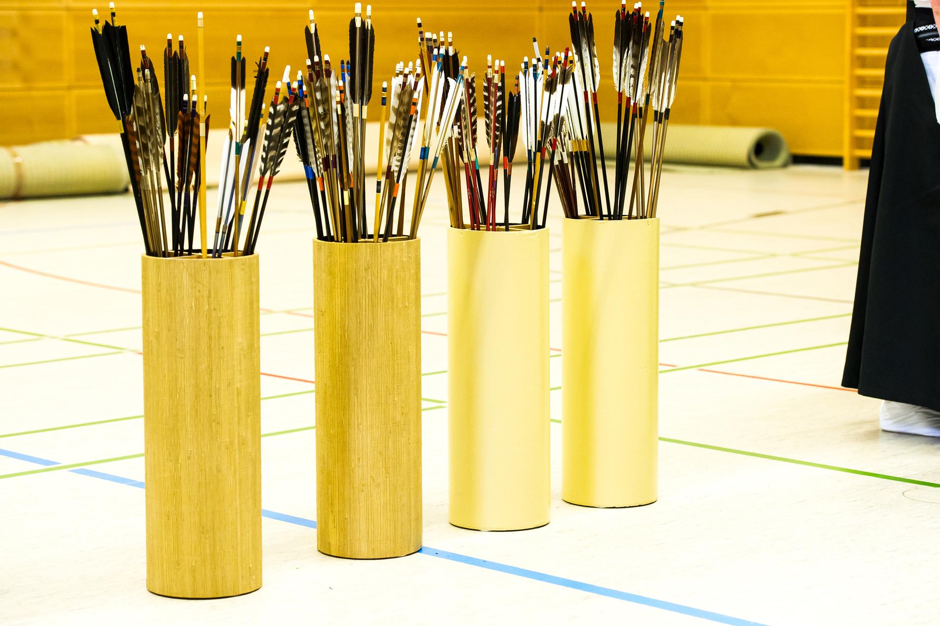 Vier Ständer mit zahlreichen Pfeilen stehen auf einem Sportboden mit bunten Linien in einer Turnhalle.