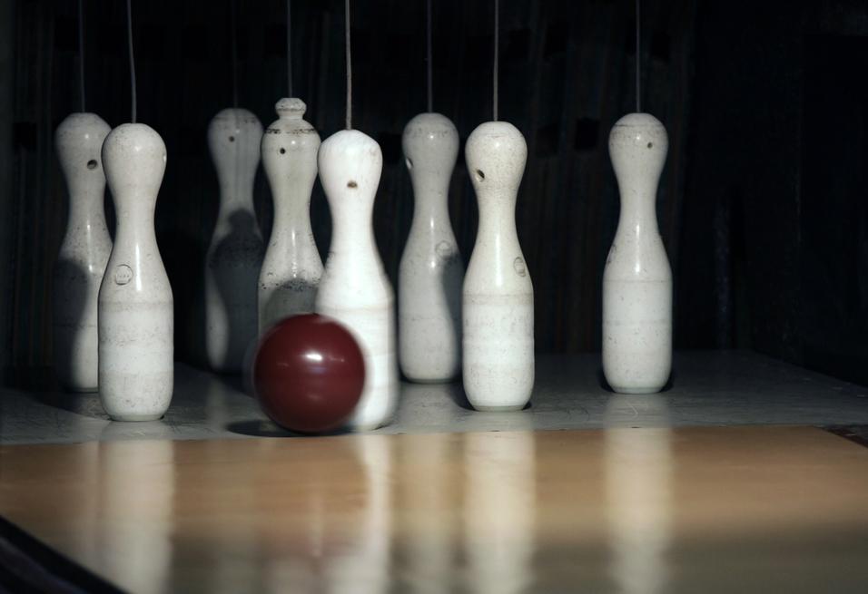 Eine rotbraune Bowlingkugel rollt auf eine Reihe weißer Kegel auf einer Bowlingbahn zu.
