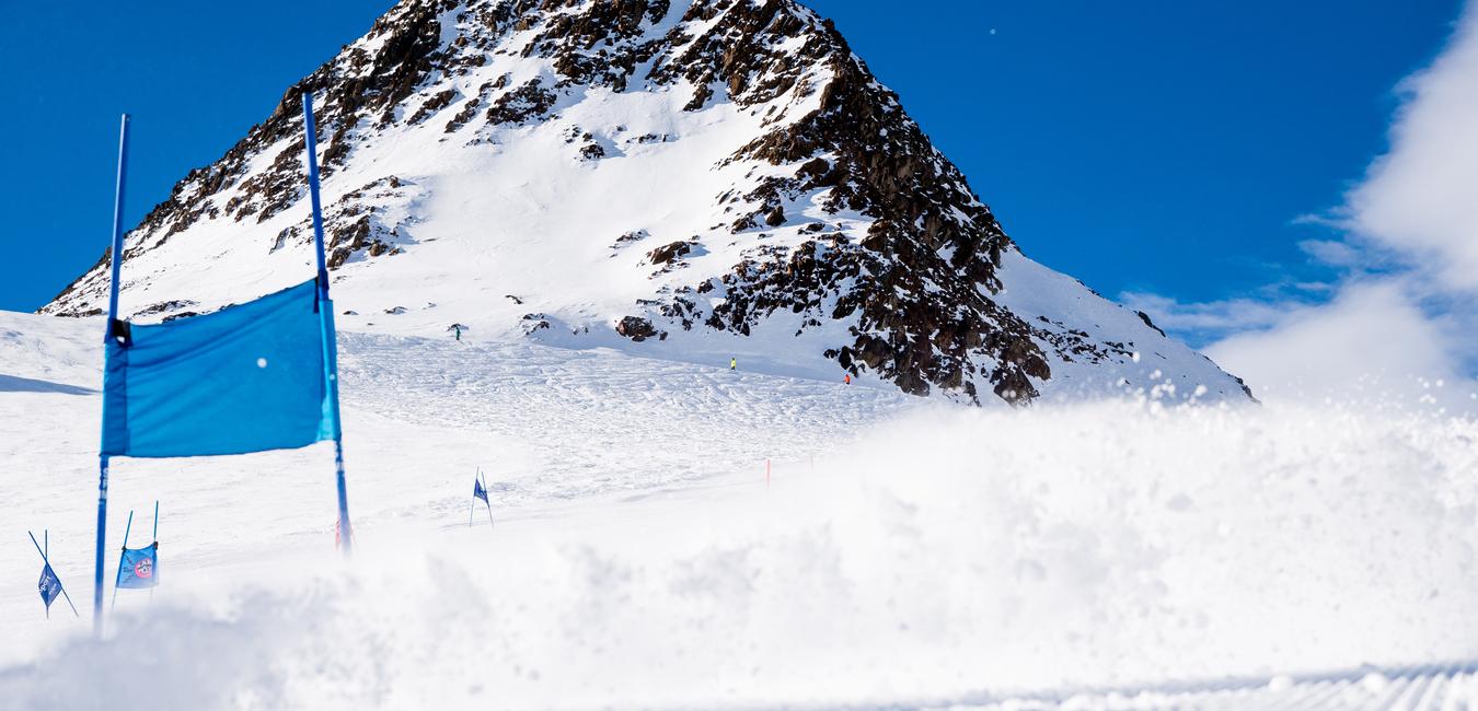 Gestreute Schneepistе mit blauen Fahnen, im Hintergrund ein schneebedeckter Berg unter blauem Himmel.