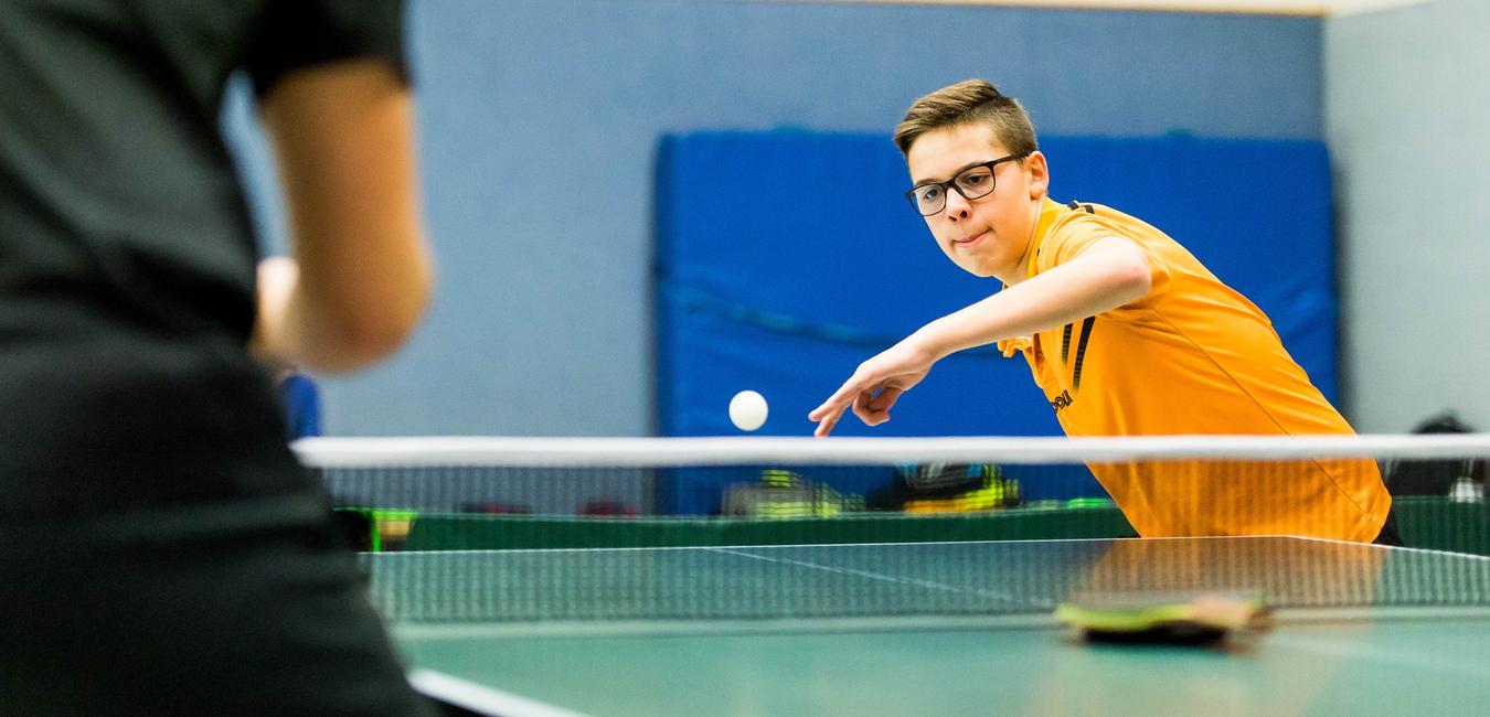 Junger Spieler in gelbem Trikot schlägt einen Tischtennisball über das Netz auf einer Tischtennisplatte.