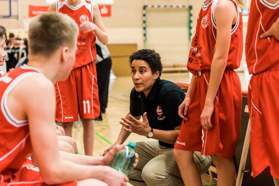 Basketballtrainerin erklärt Taktik während einer Auszeit zu Jugendlichen in roten Trikots auf dem Spielfeld.