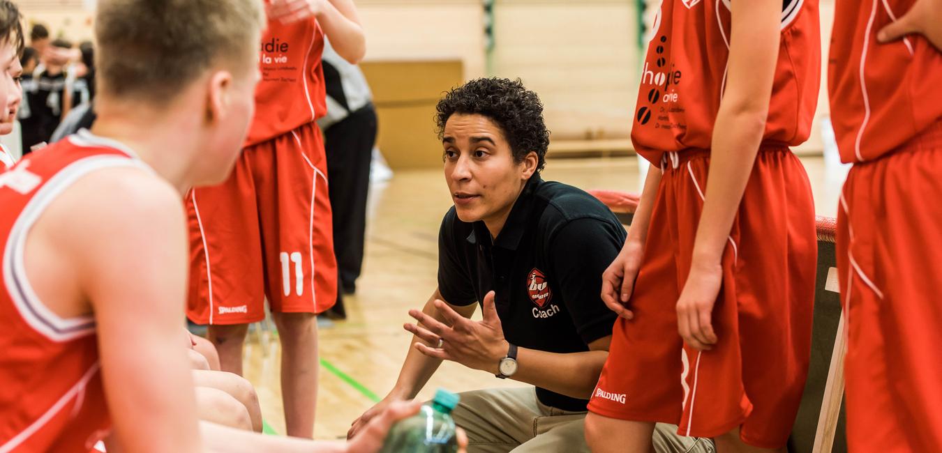 Basketballtrainerin erklärt Taktik während einer Auszeit zu Jugendlichen in roten Trikots auf dem Spielfeld.