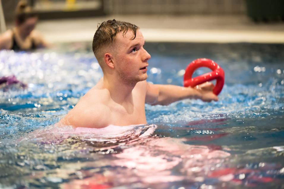 Junger Mann im Wasser hält einen roten Schwimmring und schaut aufmerksam nach oben. Hintergrund zeigt verschwommene Schwimmer.