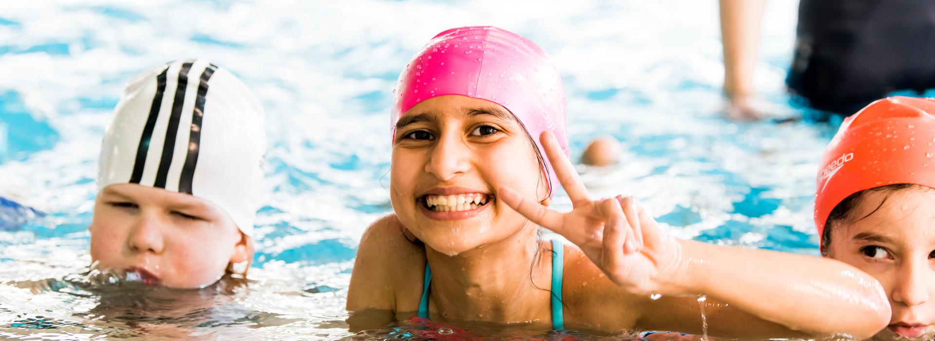 Lächelndes Mädchen mit pinker Badehaube zeigt V-Zeichen im Wasser; zwei weitere Kinder sind im Hintergrund zu sehen.